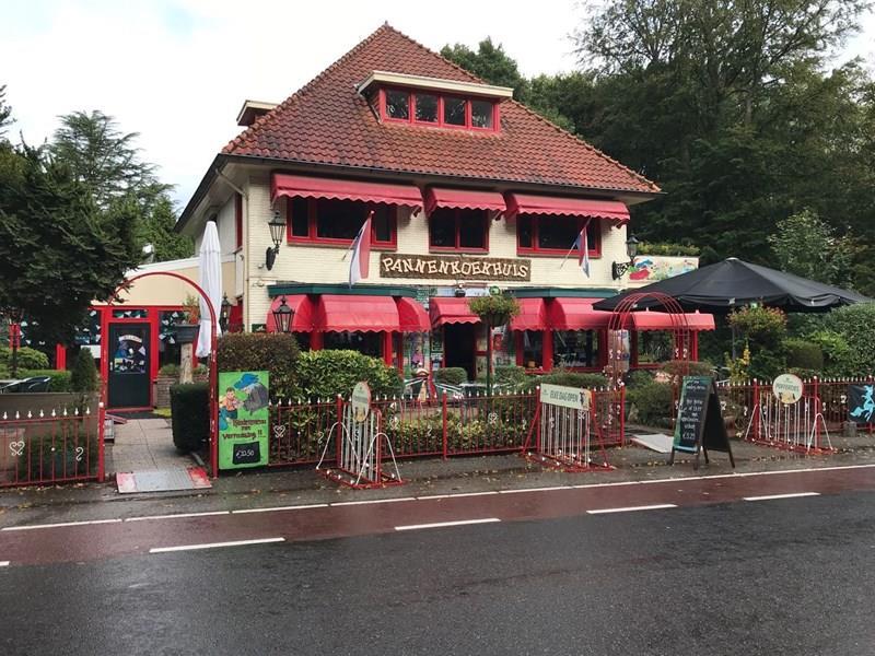 Gezellig pannenkoekenhuis met rood-witte gevel en luifels, omgeven door groen en versierde hekken aan een natte straat.